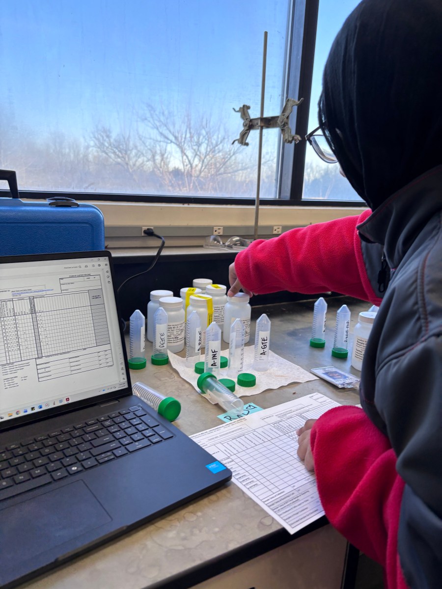 Person wearing a black hijab and a jacket with red sleeves points toward a row of labeled sample bottles and vials (including tubes marked A-GTE and A-INF) set out on a lab bench next to an open laptop showing a chain-of-custody spreadsheet, with a blue equipment case and paperwork nearby.