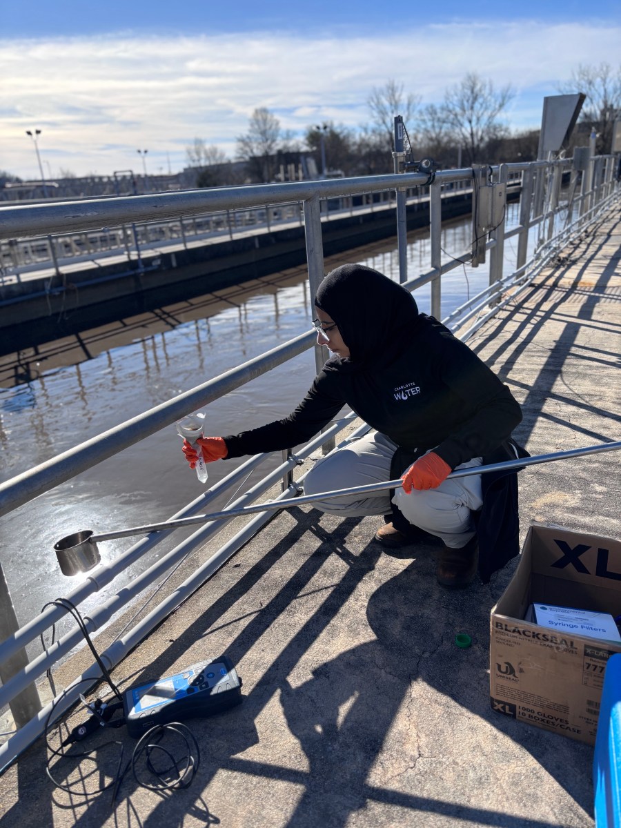 Person wearing a black hijab and a black Charlotte Water sweatshirt, wearing orange gloves and glasses, kneels at a metal railing at a wastewater treatment plant, holding a sample vial while a long metal sampling pole reaches out over brown water below a clear sky.