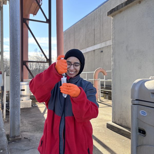 Person wearing a black hijab and a red and gray jacket, wearing orange gloves and glasses, smiles while holding a clear sample tube upright with both hands, standing at a wastewater treatment plant near a gray Hach-branded equipment cabinet and an orange pipe under a clear blue sky.