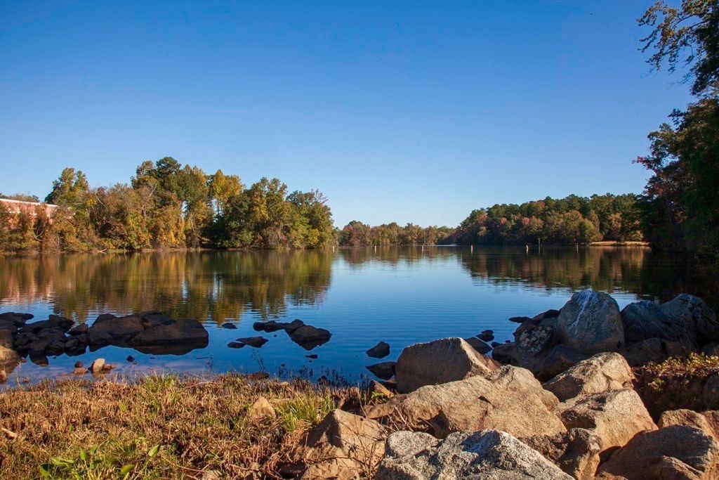 View of the lake from the shoreline with blue sky and trees in the background. The Shoreline has boulders on the right hand side.