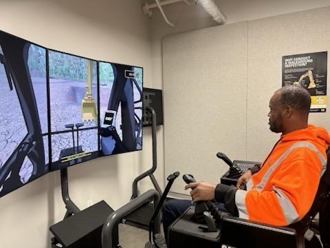A Charlotte Water employee in an orange safety hoodie uses joystick controls at an excavator simulator with three curved screens.