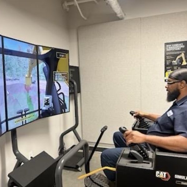A Charlotte Water employee in a navy work shirt and safety glasses operates the joystick controls of a CAT simulator, watching three curved screens that show an excavator digging at a virtual work site.